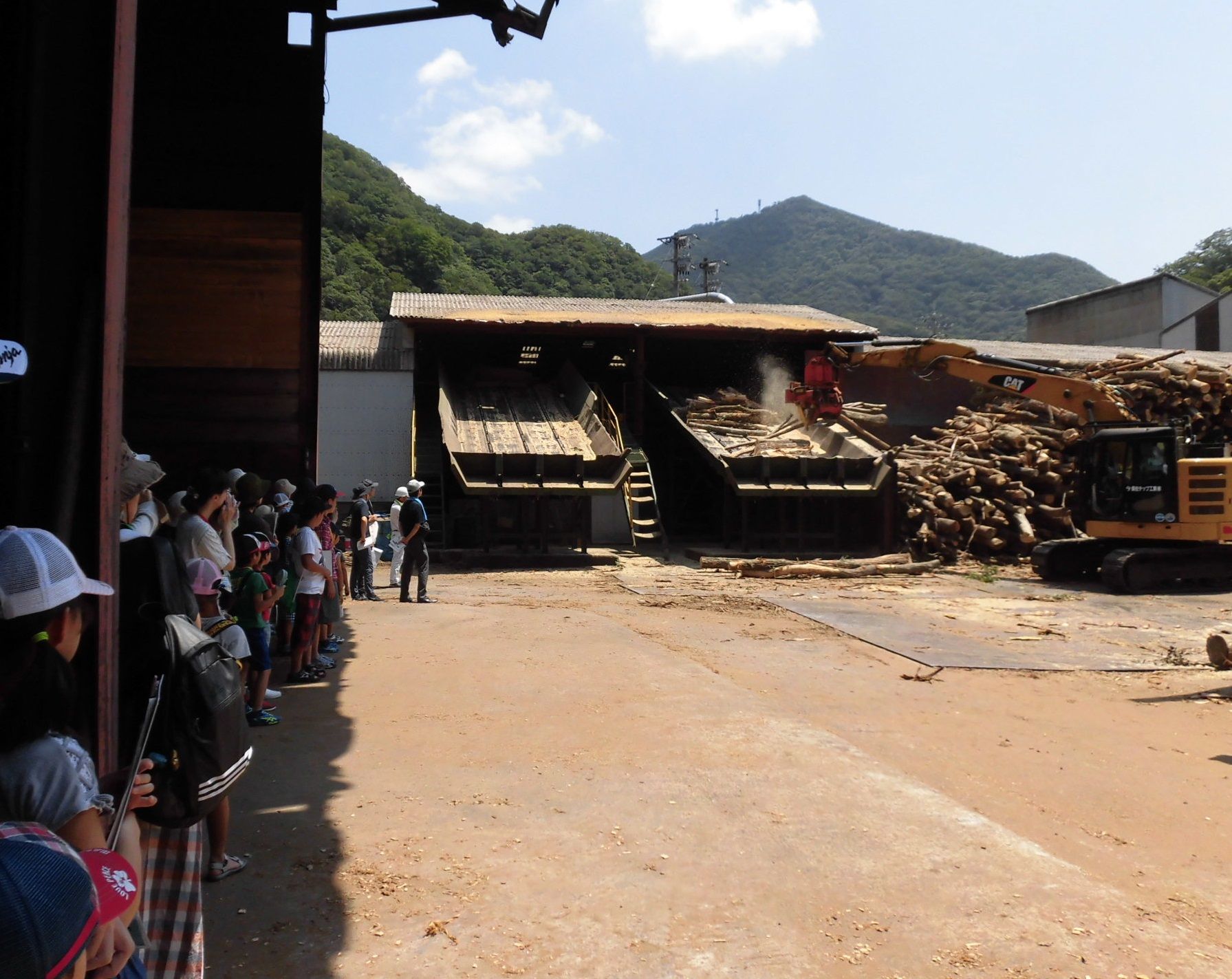 須佐チップ工業有限会社で大きな木材をショベルカーが機械に投げ込む様子を見学する受講者の写真